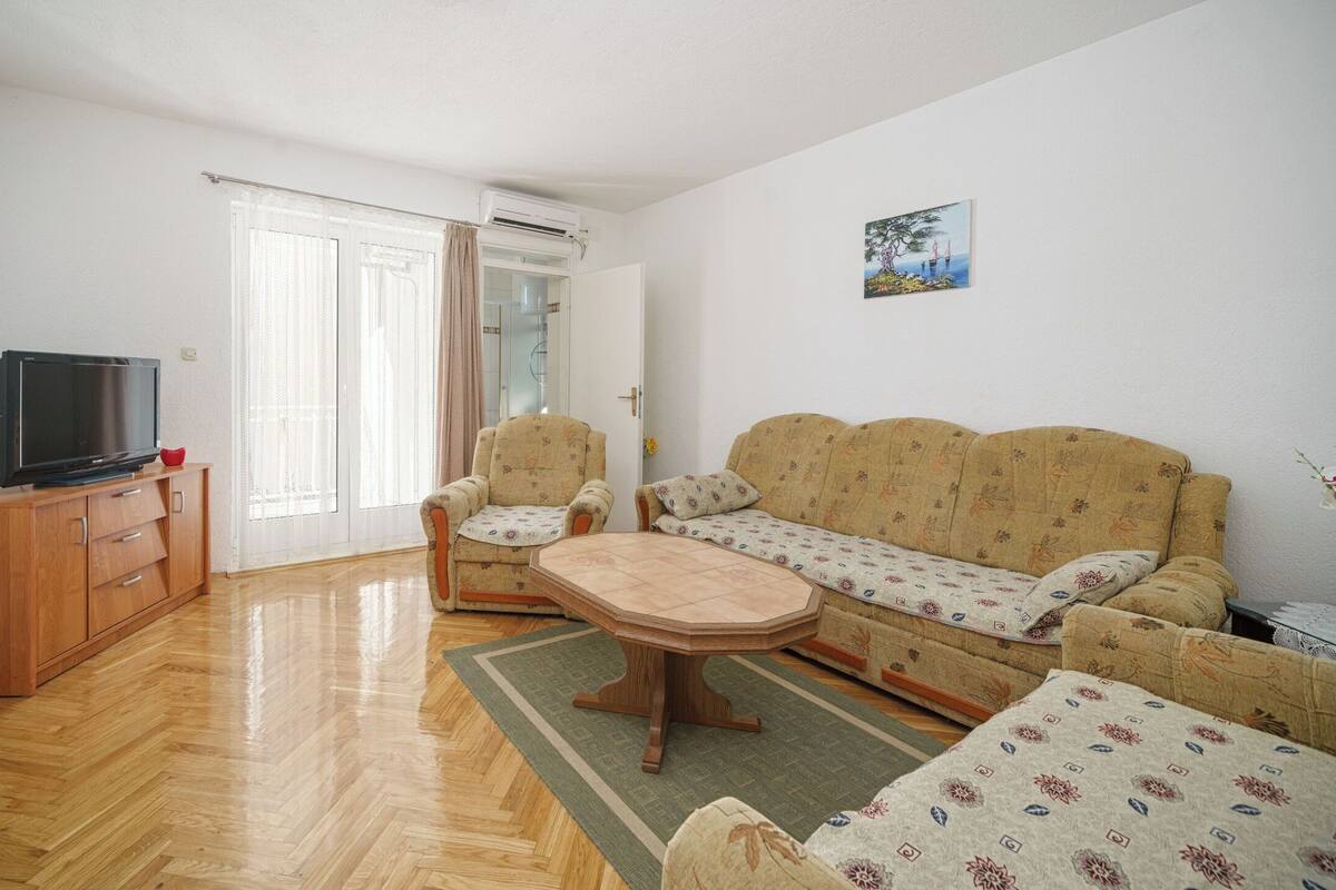 A living room with a sofa, two armchairs, a coffee table, a TV on a wooden cabinet, and a door leading to another room.