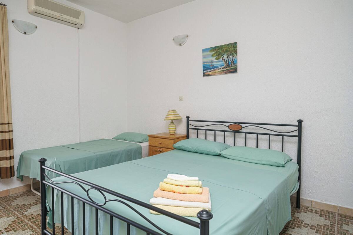 A bedroom with two single beds, a nightstand with a lamp, a painting on the wall, and an air conditioning unit.