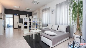 A modern living room with a sofa, a coffee table, and a bookshelf. There is a dining area in the background with a chair and a table. - House Kala (Sibenik)