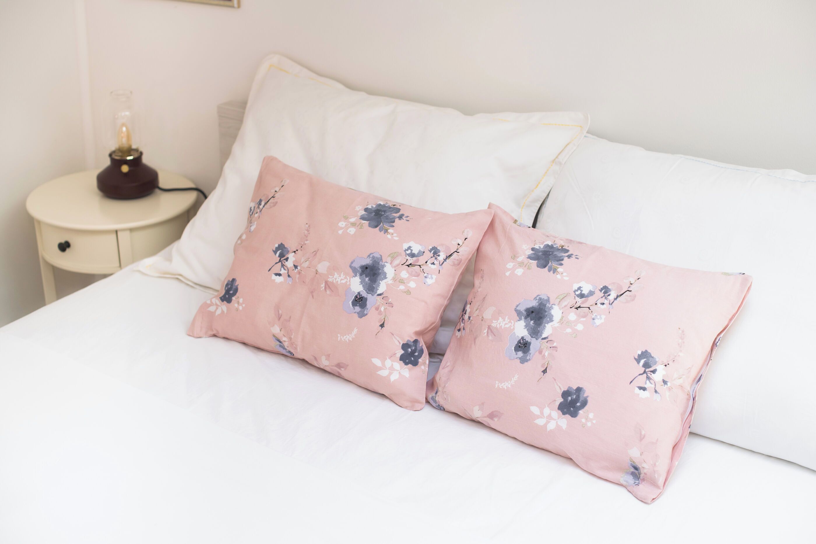 Una cama con sábanas blancas y dos almohadas con estampado floral rosa.