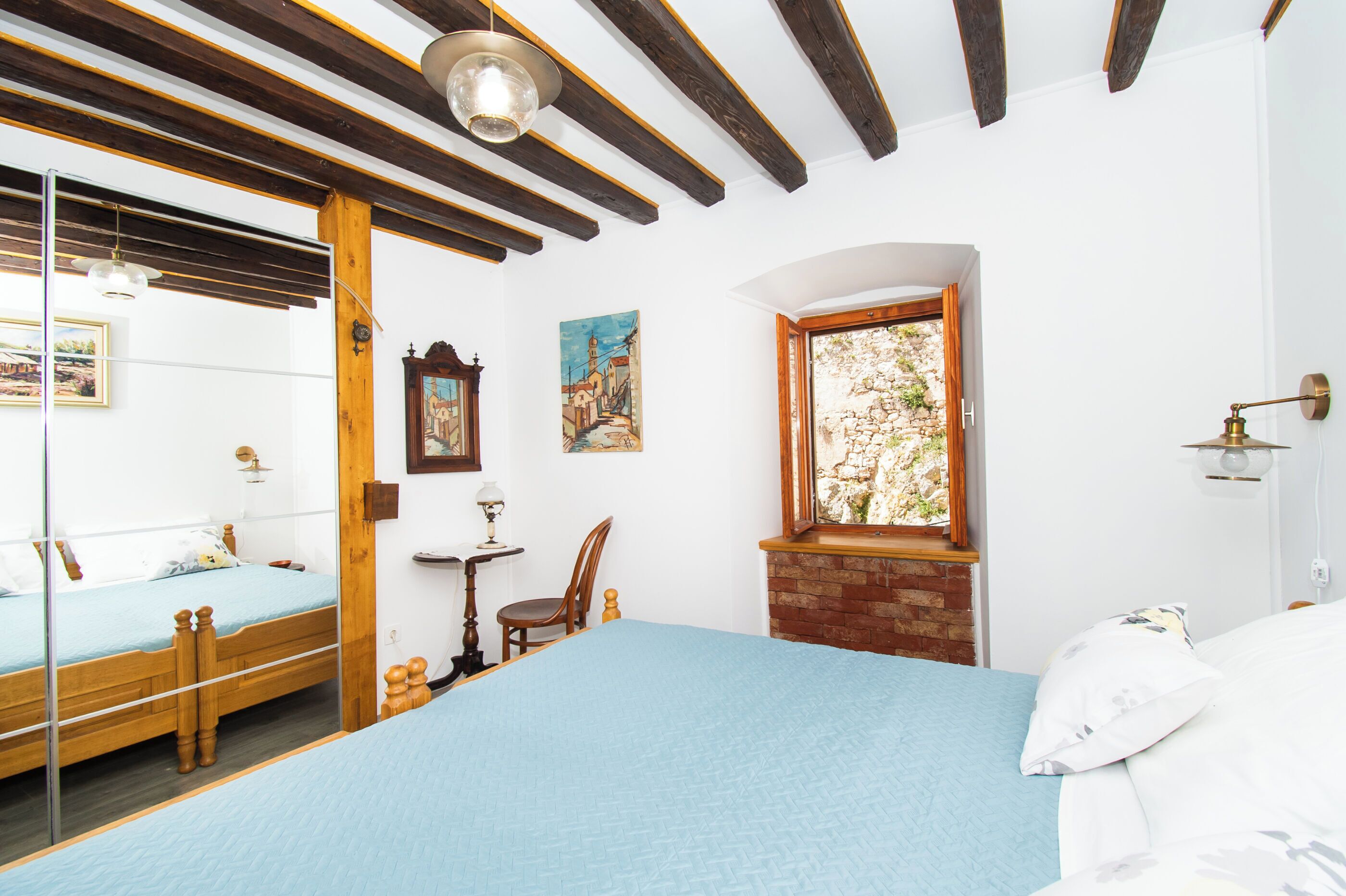 Un dormitorio con una cama, una silla, una mesita, un espejo y una ventana con vistas a la vegetación.