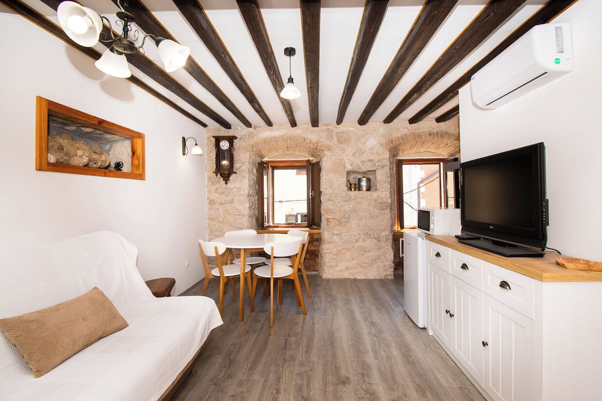 A cozy room with a white sofa, a dining table with chairs, a TV on a wooden cabinet, and a stone wall.