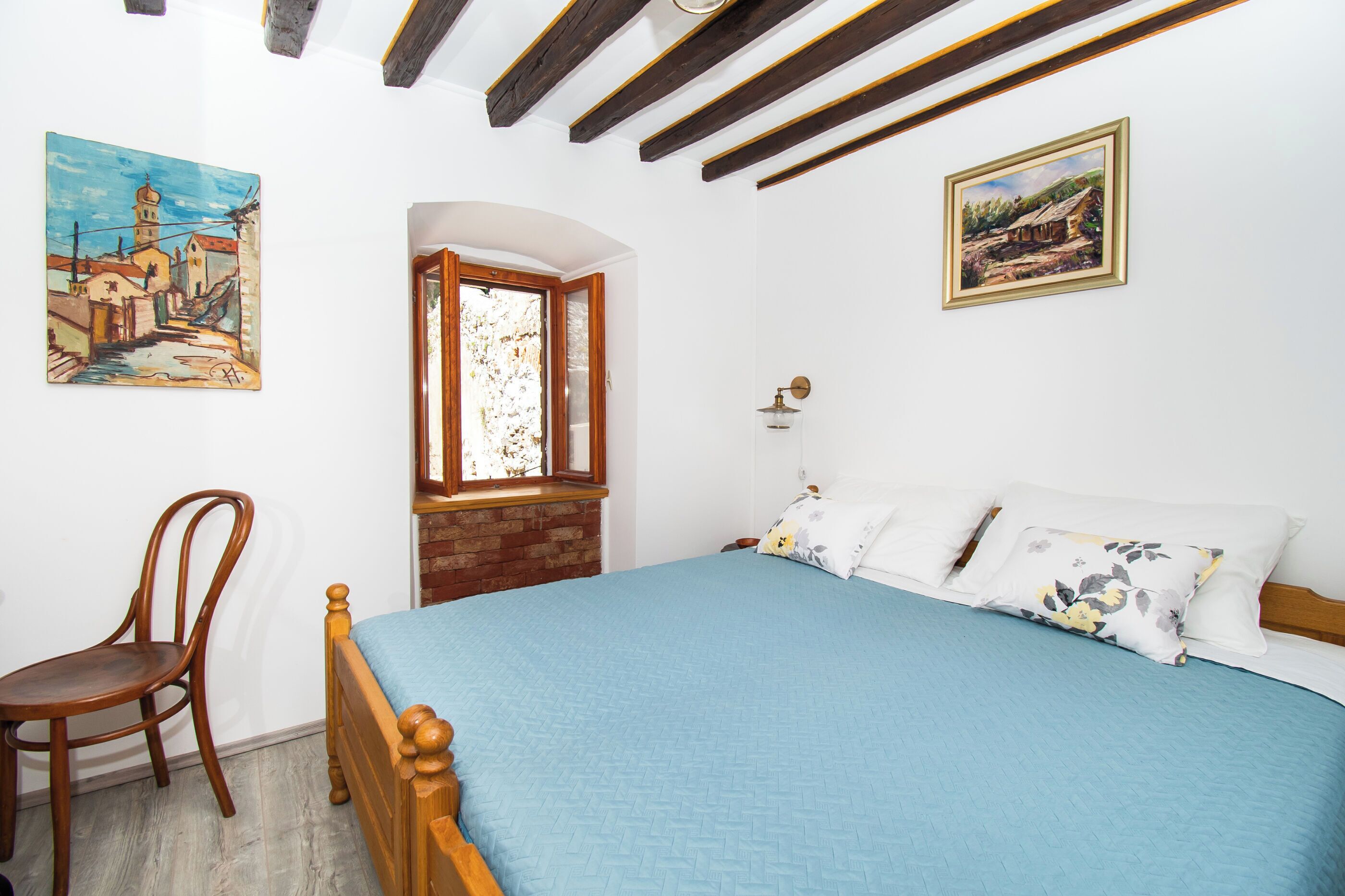 A bedroom with a wooden bed, a chair, a window, and two paintings on the wall.