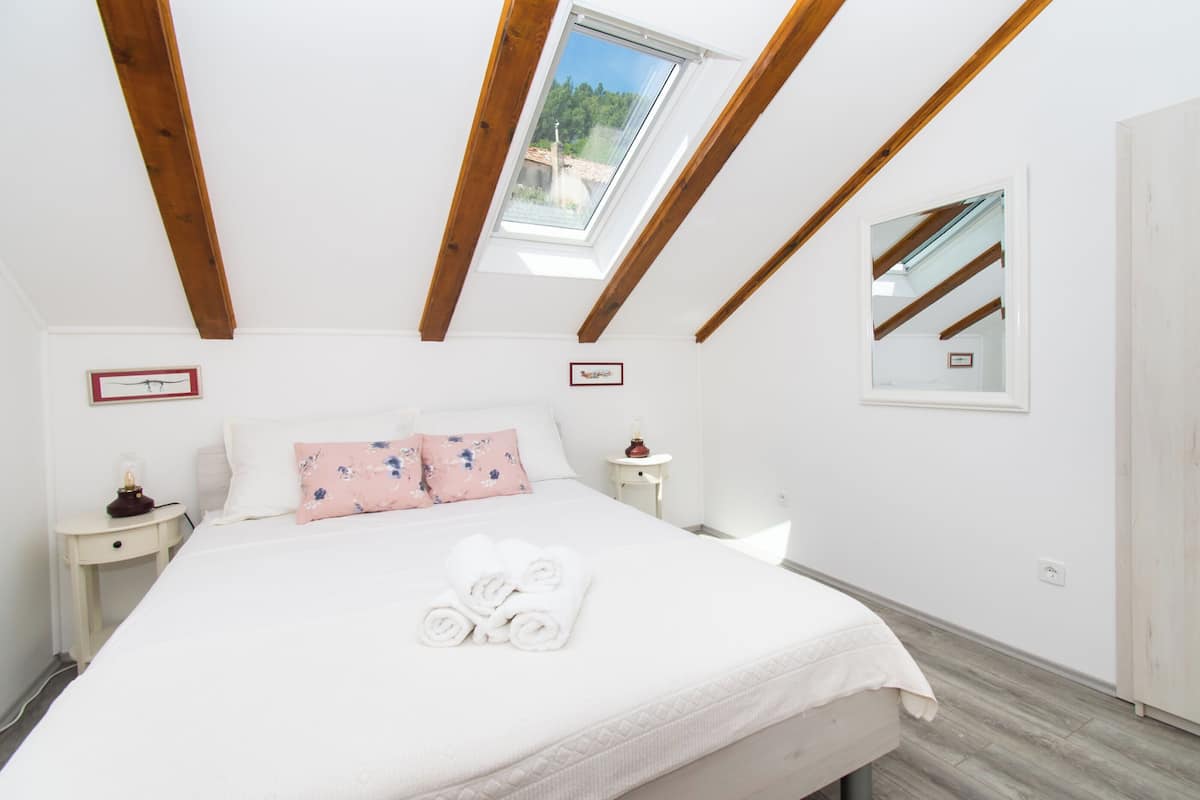 A bedroom with a skylight, a bed with white linens, a nightstand, a bedside table, and a mirror on the wall.