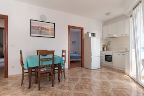A compact kitchen with white cabinets, a refrigerator, and a microwave. There is a dining area with a table and chairs, and a bedroom visible through an open door. - Apartments Anamarija (Rovinj)