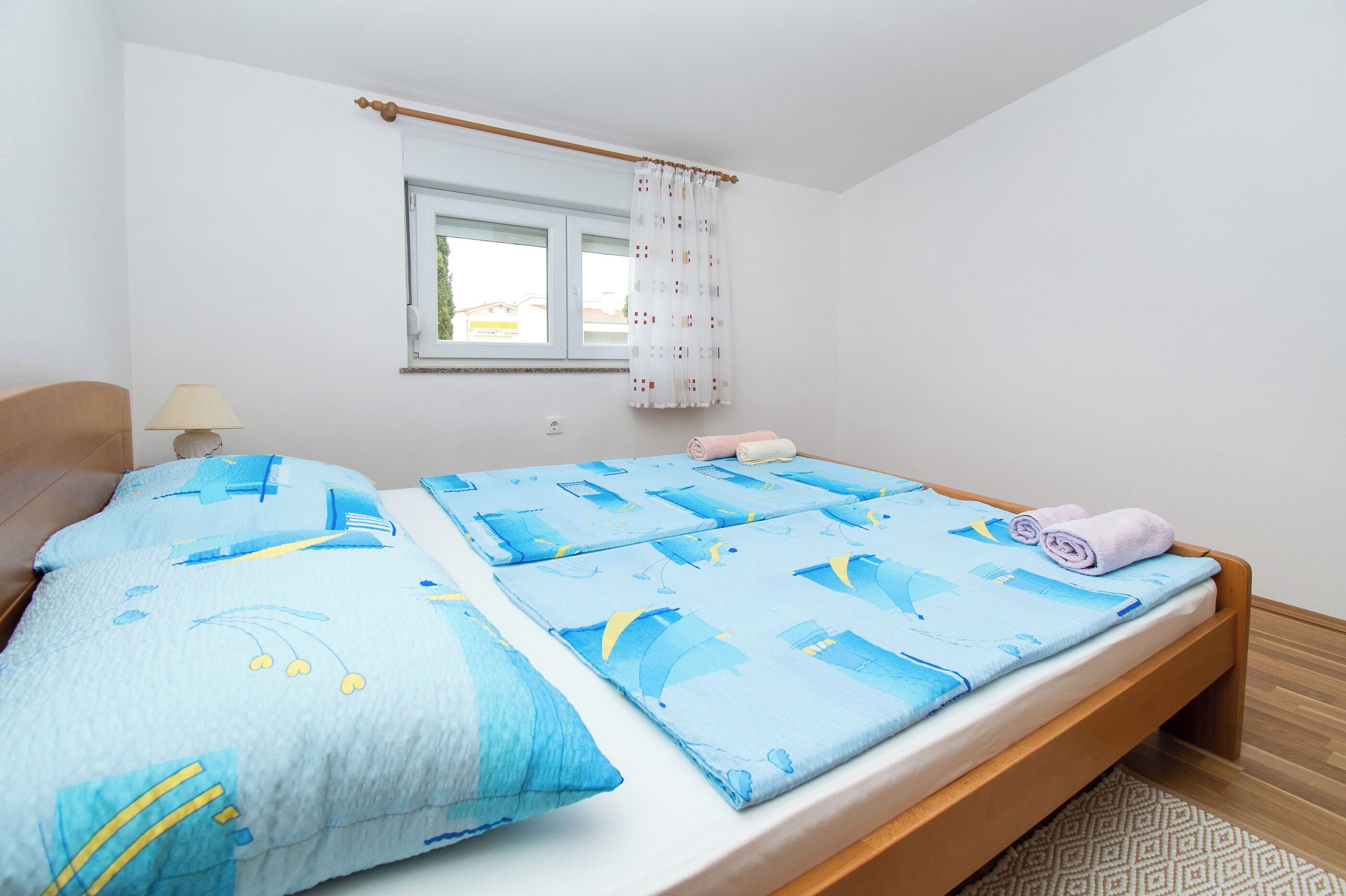 A bedroom with a wooden bed, a window with white curtains, and a bedside table with a lamp.
