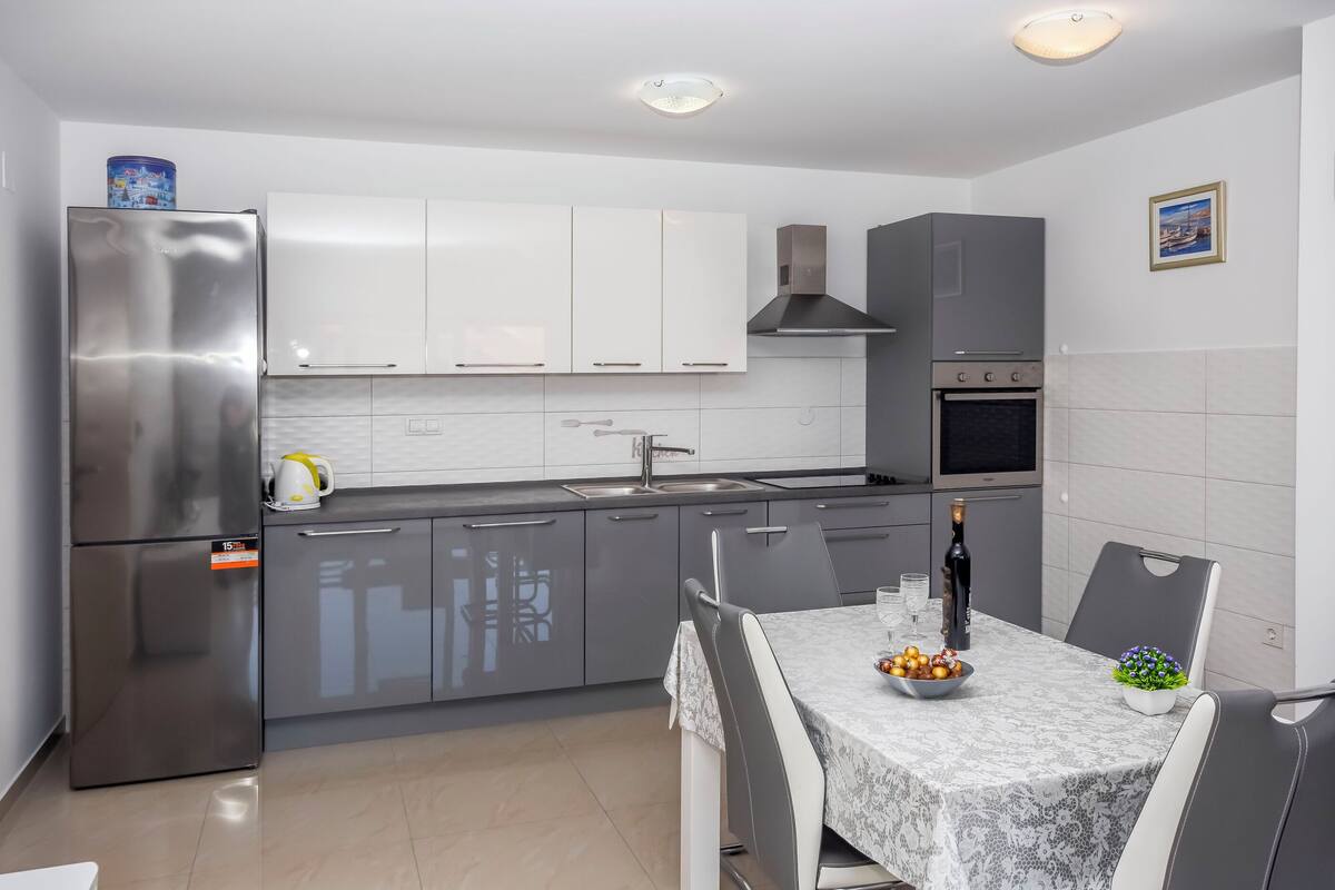 A modern kitchen with grey cabinets, a stainless steel refrigerator, and a dining area with a table set for two.