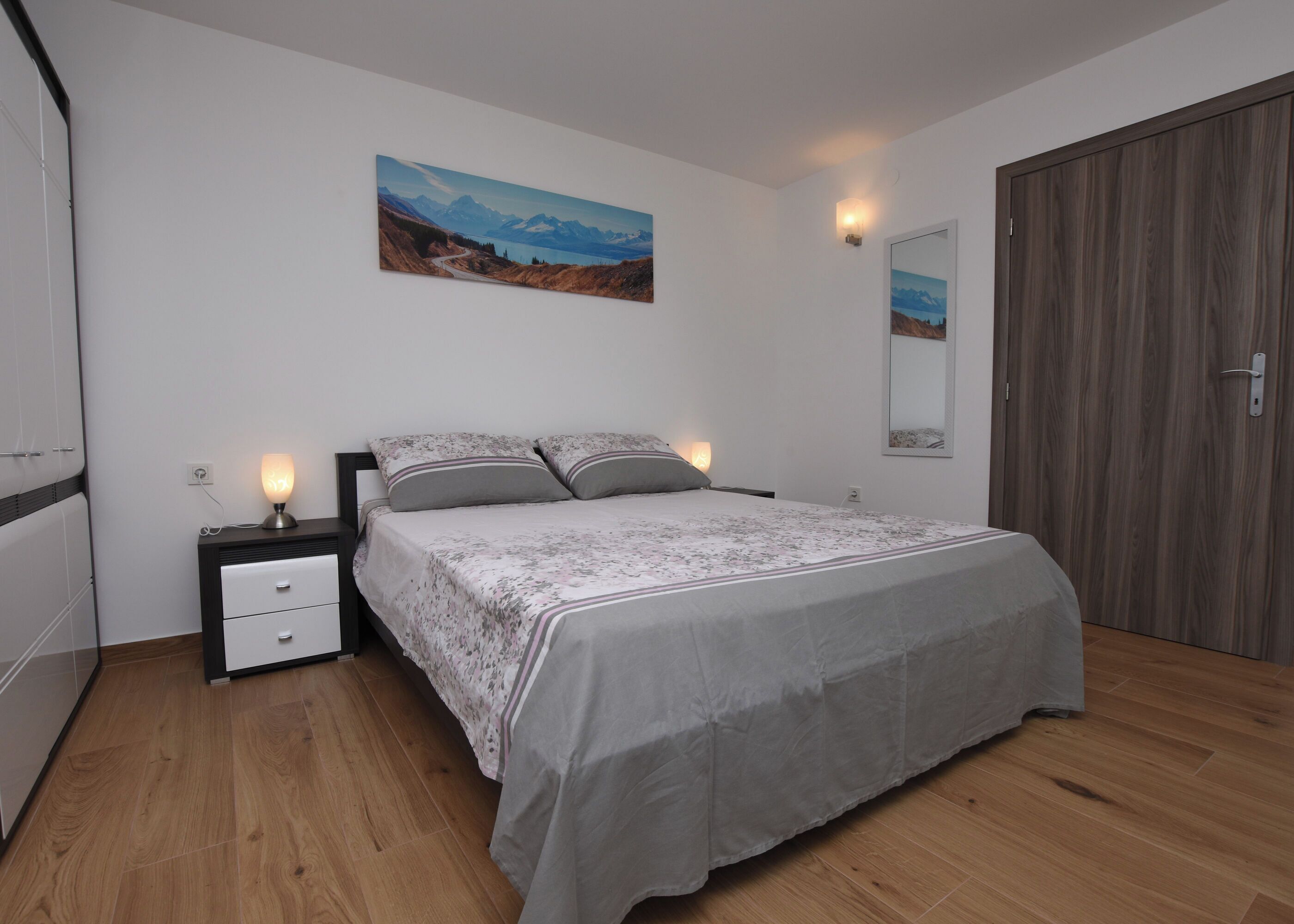 A bedroom with a bed, bedside tables, a nightstand, a wooden wardrobe, and a painting on the wall.