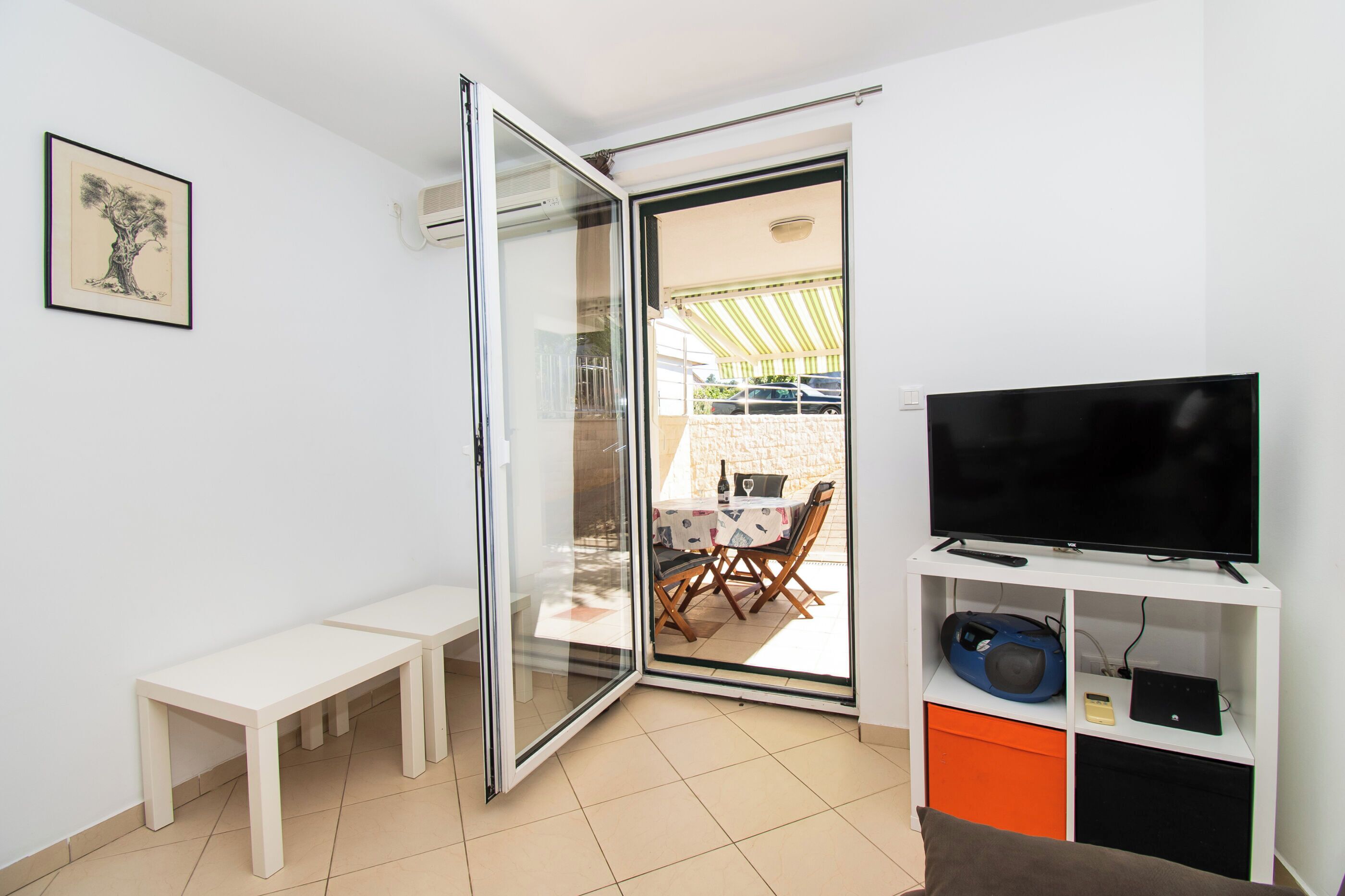 A modern living room with a sliding glass door leading to an outdoor patio, a flat-screen TV on a white stand, and a bench by the door.