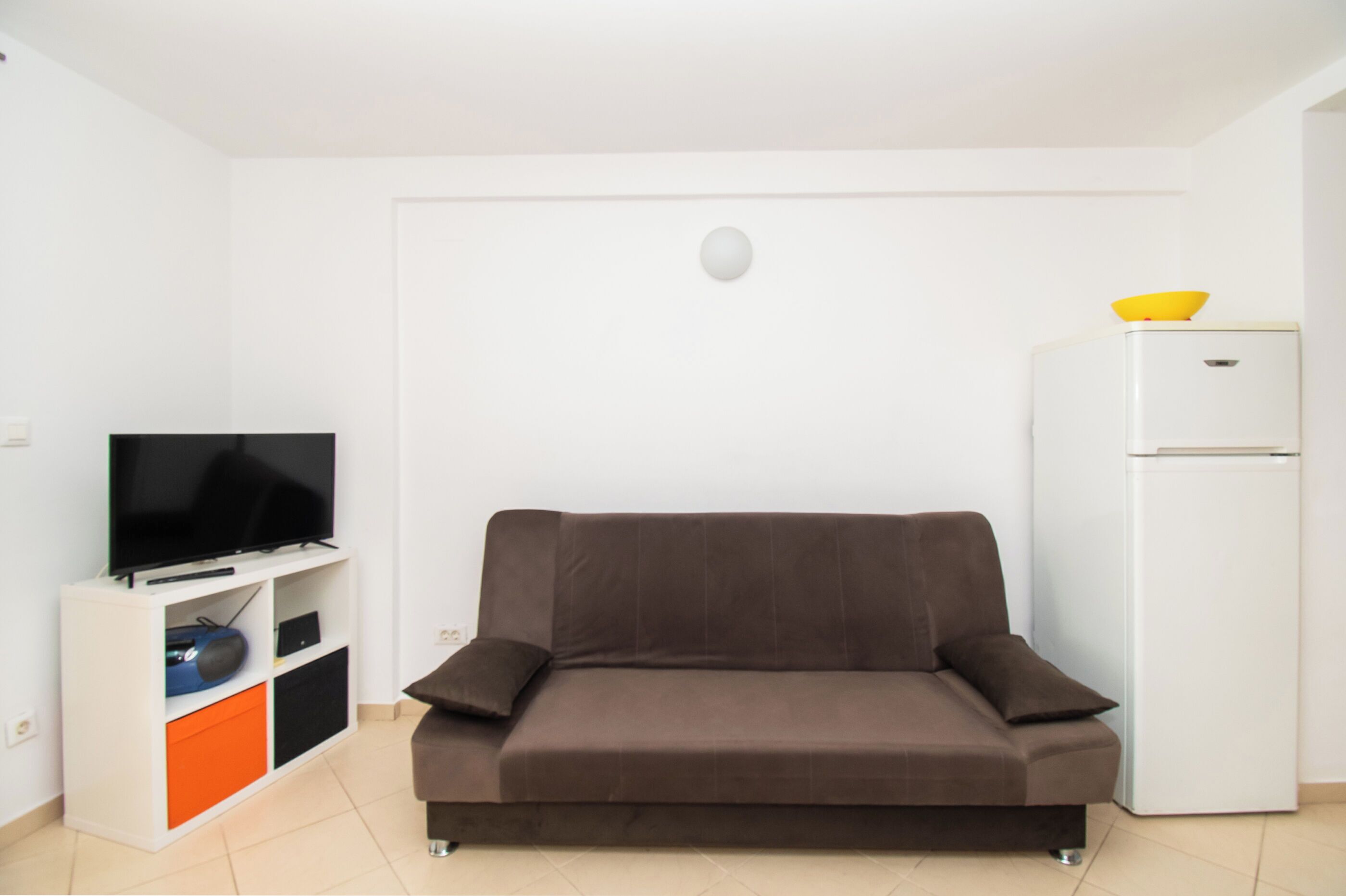 A modern living room with a brown sofa, a white entertainment unit with a flat-screen TV, and a white refrigerator.