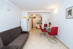 En kompakt opholdsstue med en brun sofa, et spisebord med røde stole og et lille tekøkken i baggrunden.