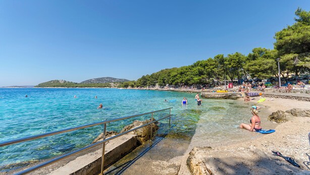Ein Strand mit klarem, blauem Wasser, Menschen beim Schwimmen und Sonnenbaden sowie einer felsigen Küste.