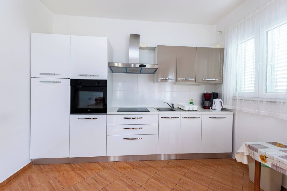 Une cuisine moderne avec des armoires blanches, une hotte de cuisinière en acier inoxydable, un four encastré, un évier et une fenêtre avec des stores.