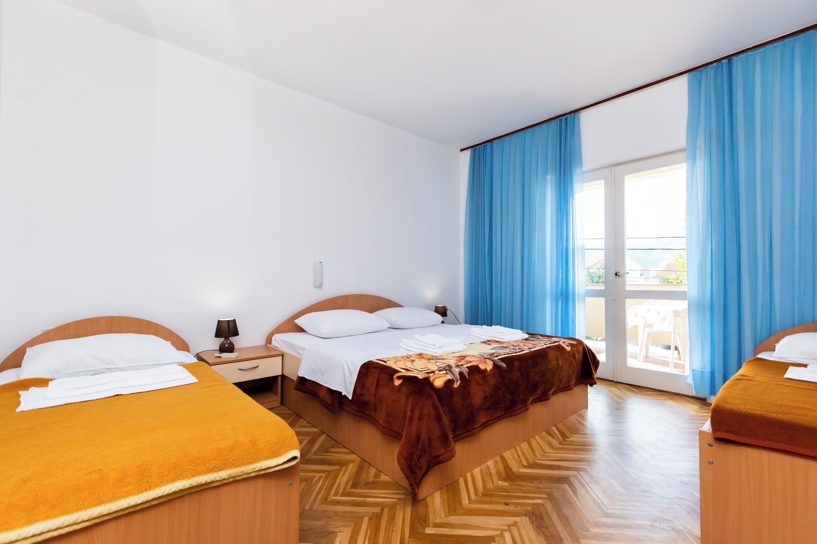 A hotel room with two single beds, a wooden floor, a large window with blue curtains, and a balcony.