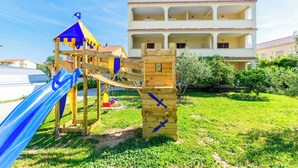 A playground with a slide and climbing structures, a multi-story building with balconies, and a clear sky. - Apartments Ivan i Marija (Lopar)