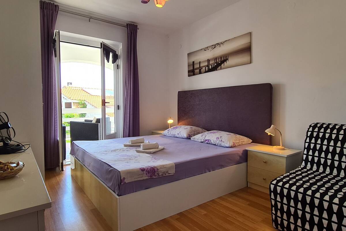 A bedroom with a bed, bedside table, lamp, and a view of the outside.