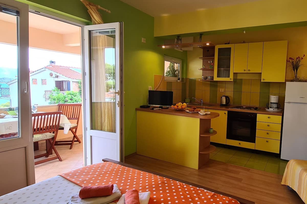 A compact room with a bed, a sofa, a small kitchen, and a balcony with a view of houses and the sea.