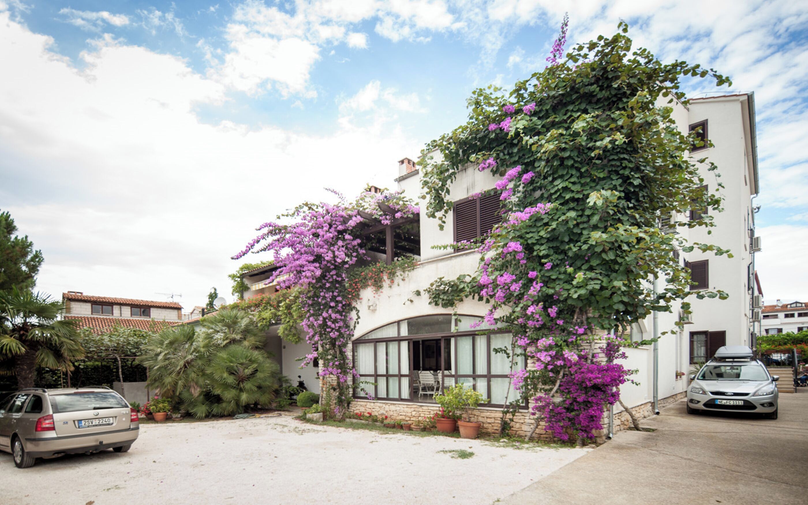Un bâtiment à deux étages, doté d’une façade blanche et recouvert de plantes grimpantes aux fleurs violettes. Une voiture est garée devant, et une autre est stationnée dans la rue.