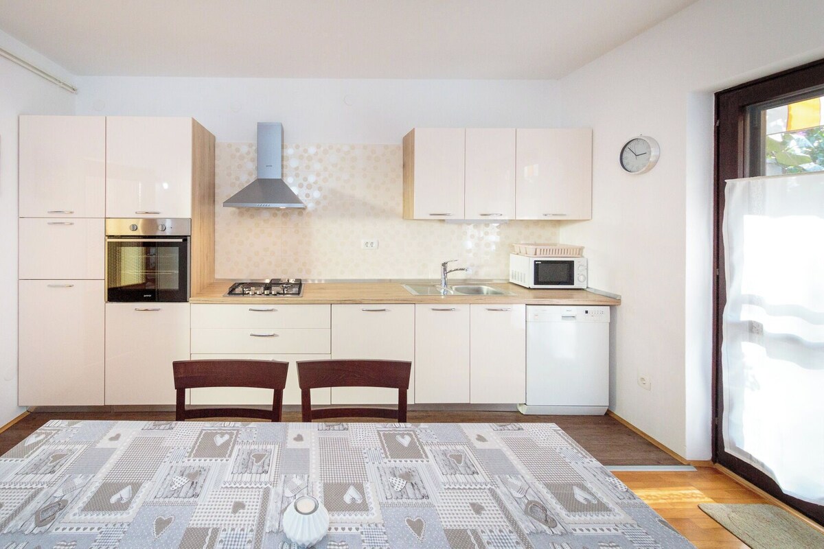 A compact kitchen with white cabinets, a built-in oven, a microwave, and a sink. There is a dining table with two chairs and a patterned rug.