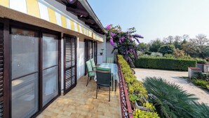 Eine Terrasse mit Tisch und Stühlen, ein Garten mit Pflanzen und einer Hecke, sowie ein Gebäude mit großen Fenstern und Fensterläden.