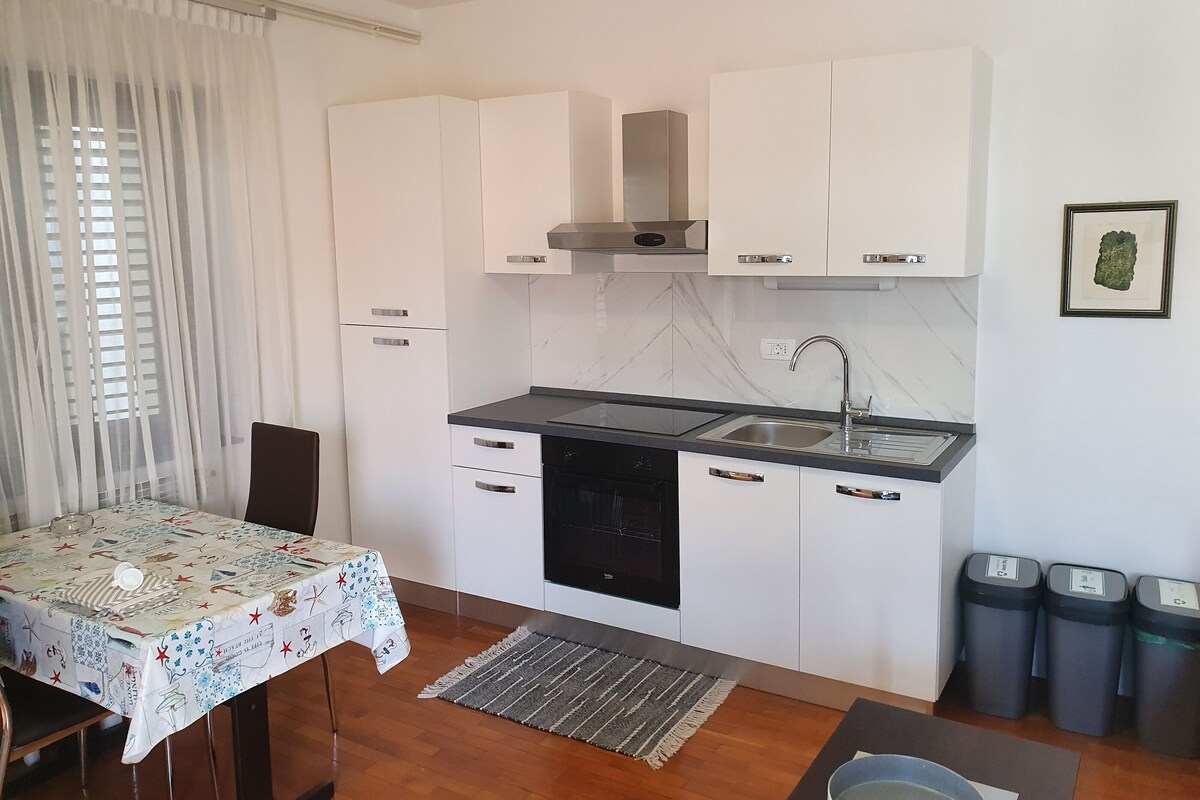 A compact kitchen with white cabinets, a black countertop, and a built-in stove. There is a dining table with a patterned tablecloth and a rug on the floor.