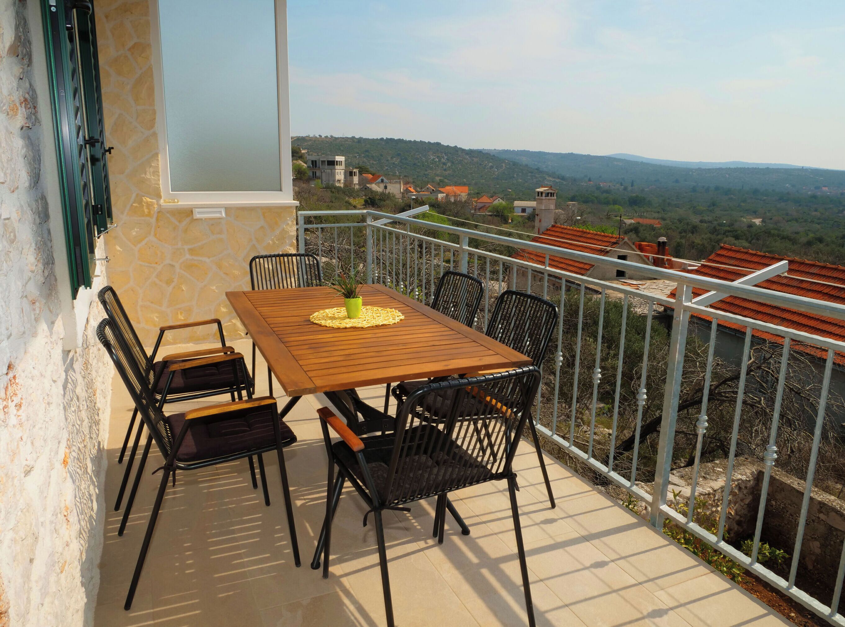 A balcony with a wooden table and chairs, a potted plant, and a view of a hilly landscape with buildings.