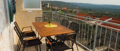 A balcony with a wooden table and chairs, a potted plant, and a view of a hilly landscape with buildings.