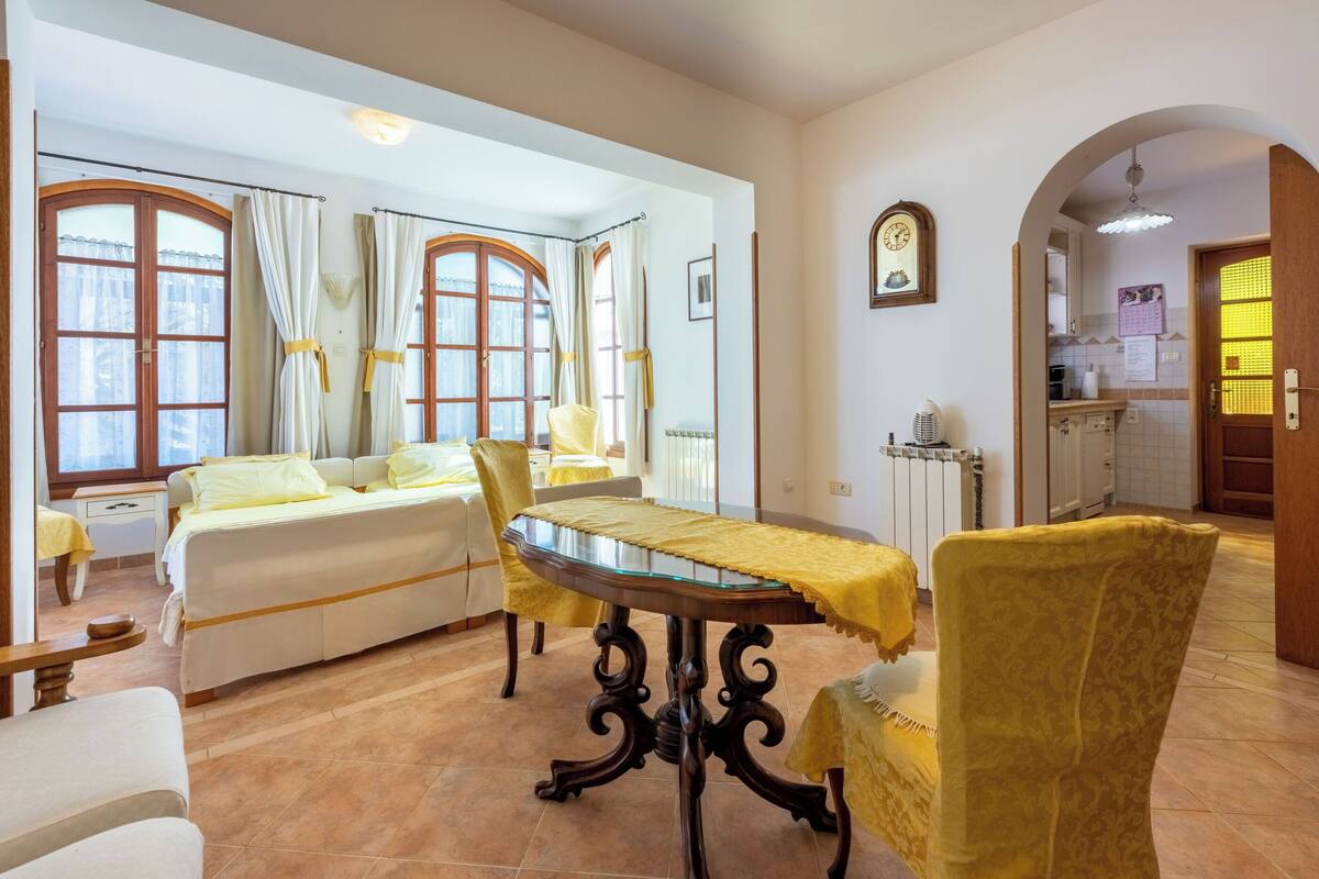 Une pièce comprenant un lit, une table à manger avec des chaises jaunes, un coin cuisine et une salle de bain visible à travers une arche.
