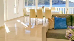 A spacious living room with a large window offering a view of a body of water and hills. There is a dining area with chairs and a table, and a sofa with cushions. - Apartment Sammy (Sali)