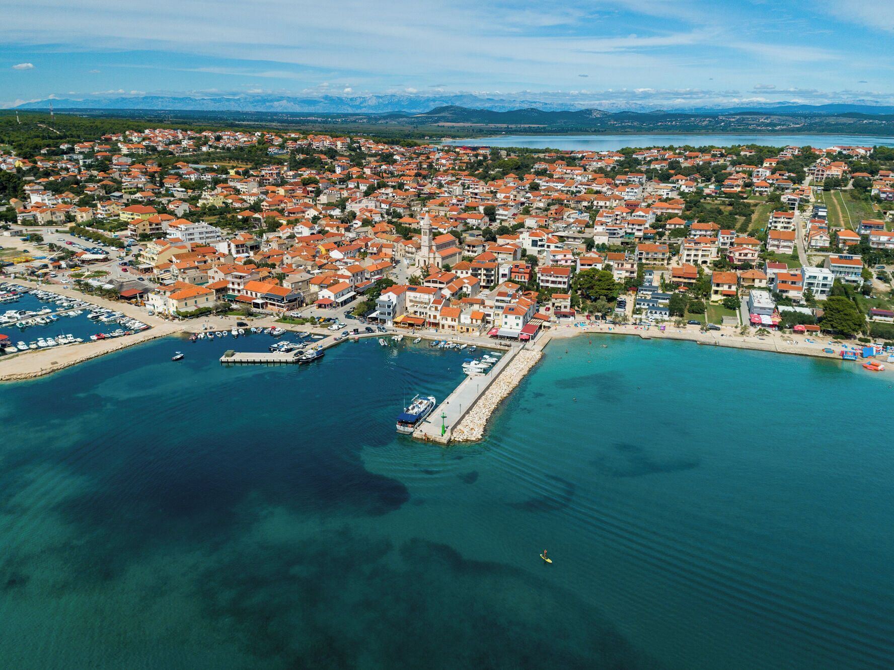 An aerial view of a coastal town with a marina, clear blue waters, and a mountainous backdrop.