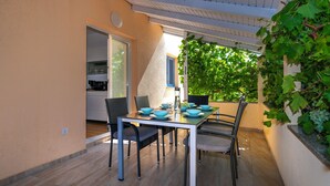 Un área de comedor al aire libre con una mesa preparada para cuatro personas, rodeada de sillas, y una cocina visible a través de una puerta abierta.
