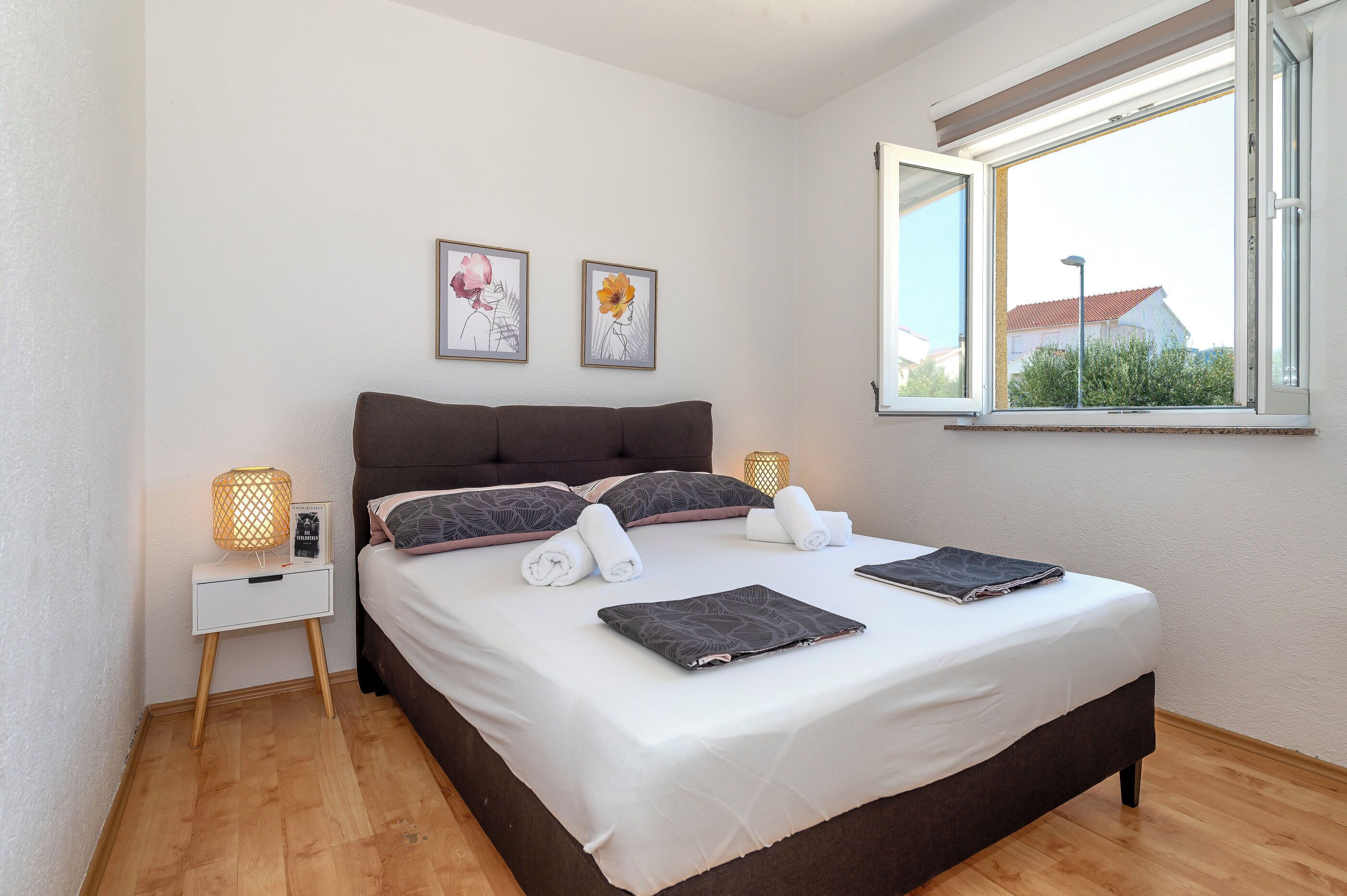 A neatly made bed with white and dark bedding, two framed artworks on the wall, and a window with a view of a building and greenery outside.
