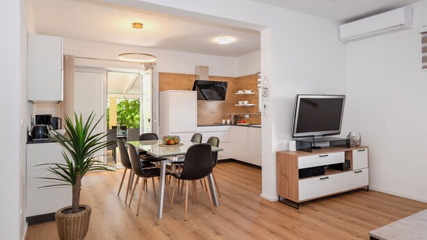 Una cocina moderna con armarios blancos, encimera de madera y horno empotrado. Hay un comedor con mesa y sillas, una planta en maceta y un televisor sobre un mueble de madera.