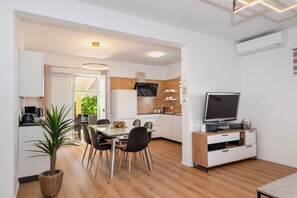 A modern kitchen with white cabinets, a wooden countertop, and a built-in oven. There is a dining area with a table and chairs, a potted plant, and a television on a wooden stand. - Apartment Dora Family Only (Vodice)