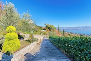 Ein gepflasterter Weg, gesäumt von Grünpflanzen und Steinmauern, führt zu einer Aussichtsplattform mit Blick auf das Meer und die entfernten Hügel.