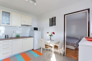 A compact kitchen with white cabinets, a microwave, and a refrigerator. There is a dining area with a table and chairs, and a bed with a patterned blanket. - House Cvetko (Lovran)
