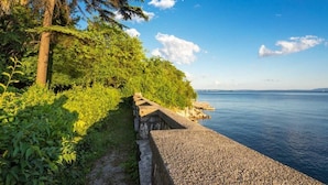 A coastal walkway with a stone railing, lush greenery, and a clear view of the sea. - Apartment Royal Studio (Rijeka)
