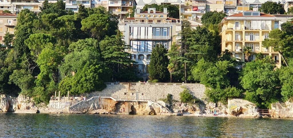 A coastal cityscape with buildings, a stone bridge, and dense greenery.