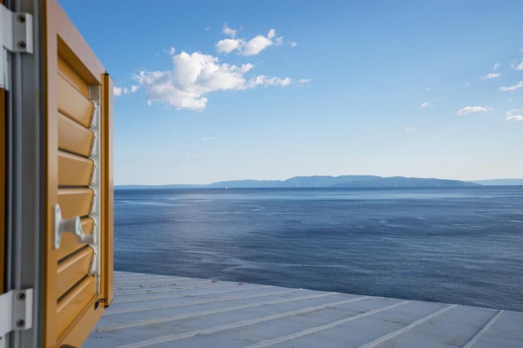 Vista desde una ventana con marco metálico y puerta de madera, que da a un mar tranquilo con formas de tierra a lo lejos.
