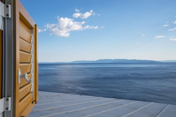 Une vue depuis une fenêtre dotée d’un cadre en métal et d’une porte en bois, donnant sur une mer calme et sur des reliefs lointains.