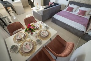A modern hotel room with a dining area set for four, a grey sofa, a bed with pink pillows, and a small kitchen area. - Apartment Royal Studio (Rijeka)