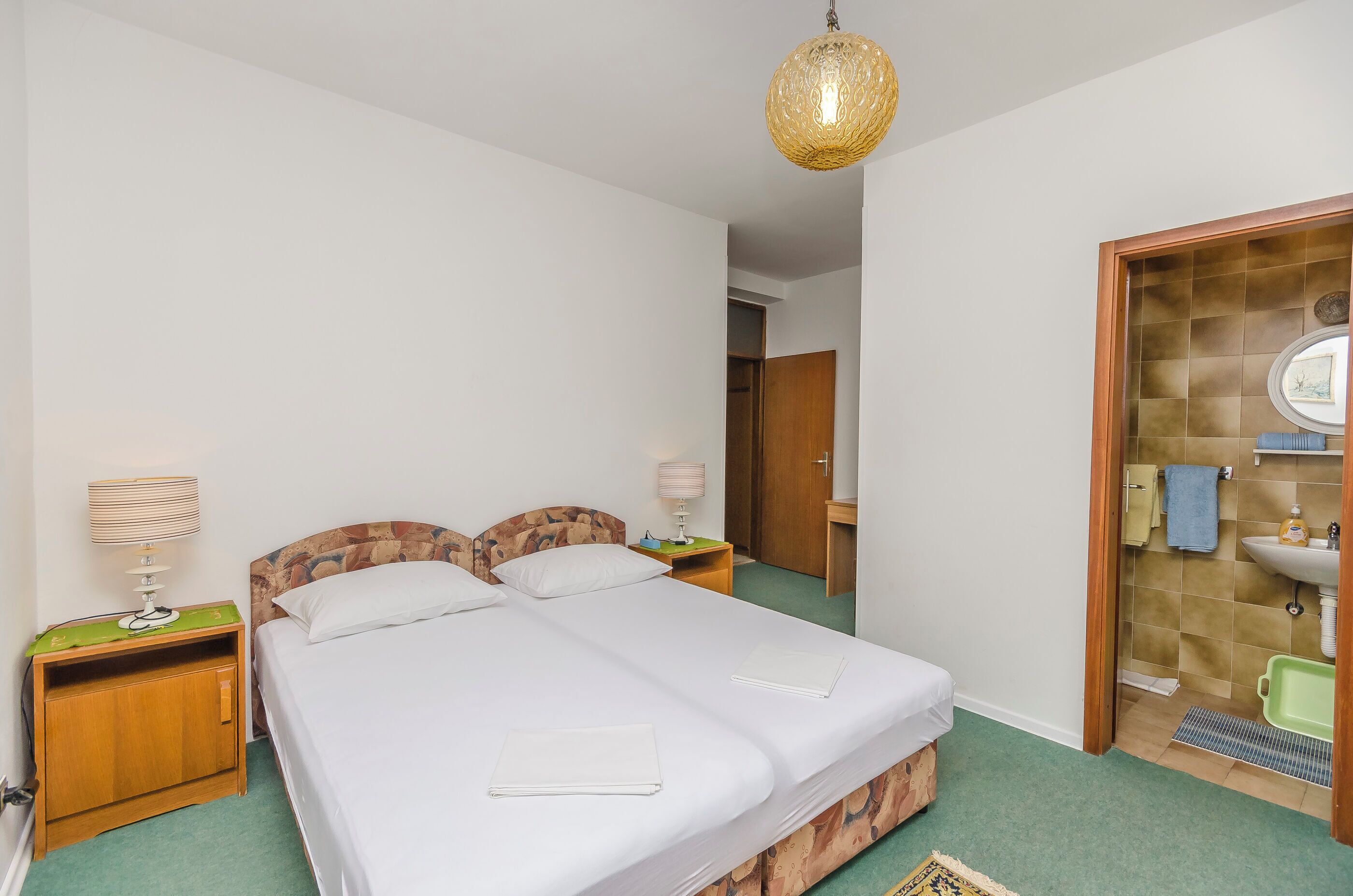 A bedroom with two beds, a wooden nightstand with a lamp, a green carpet, and a bathroom with a sink and mirror.