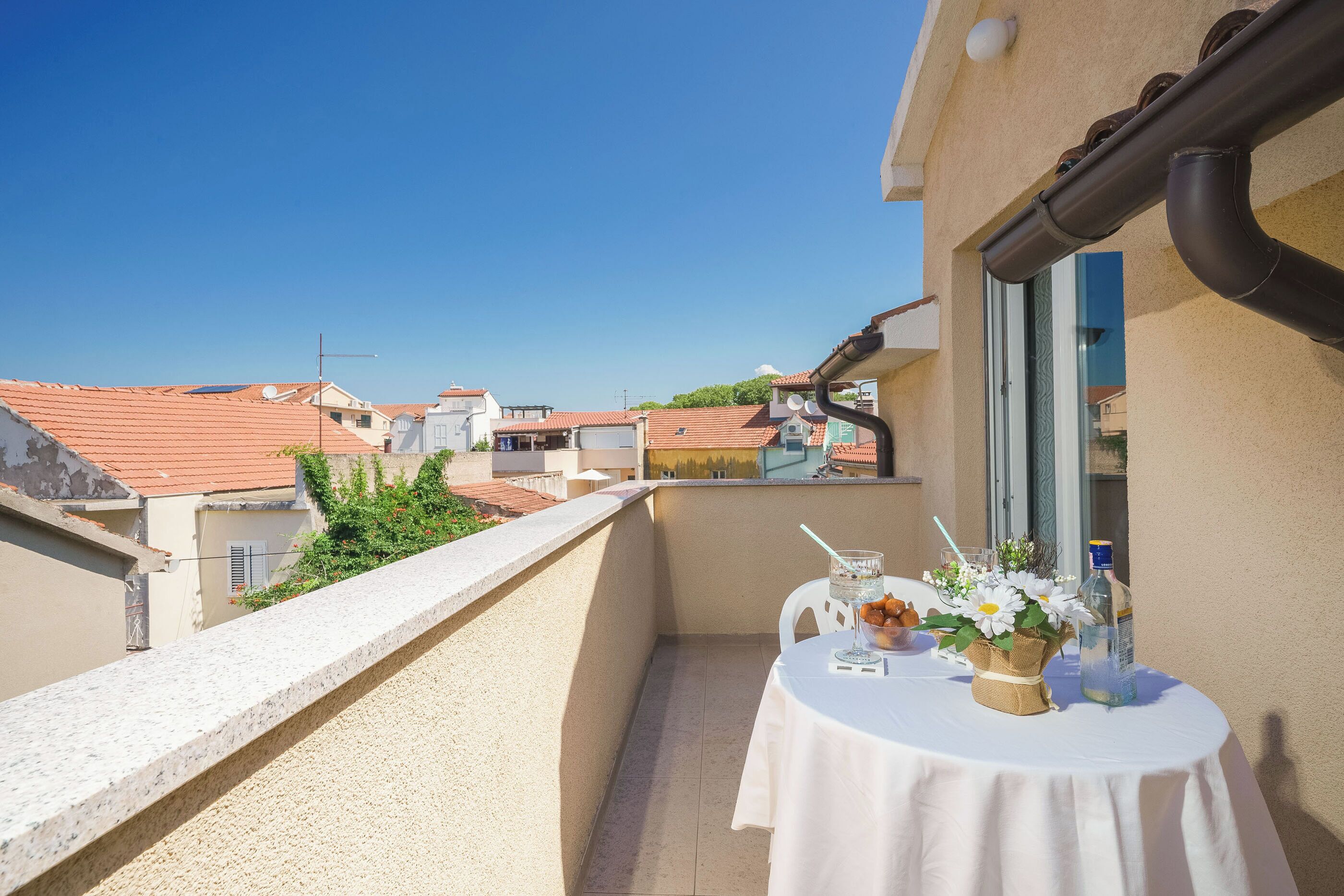 A balcony with a table set for two, featuring a vase with flowers, a bottle, and a glass.