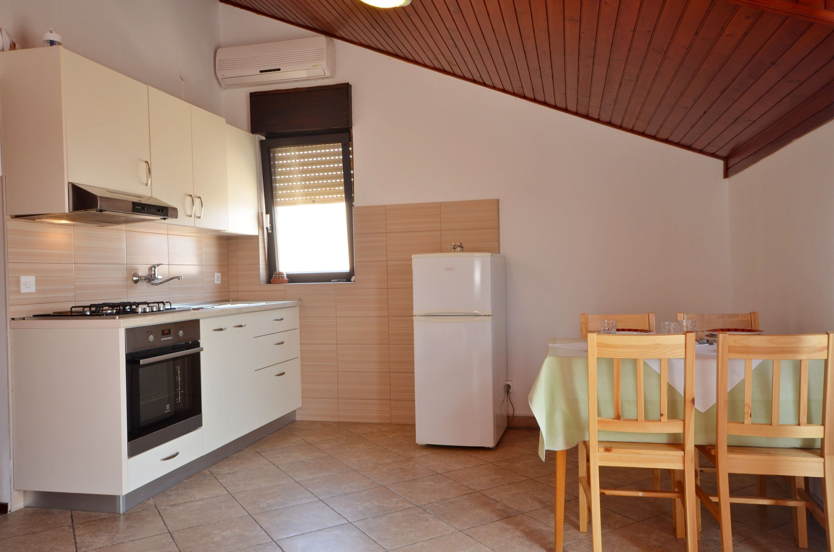 A compact kitchen with white cabinets, a stove, oven, and a refrigerator. There is a dining table with chairs and a window with blinds.