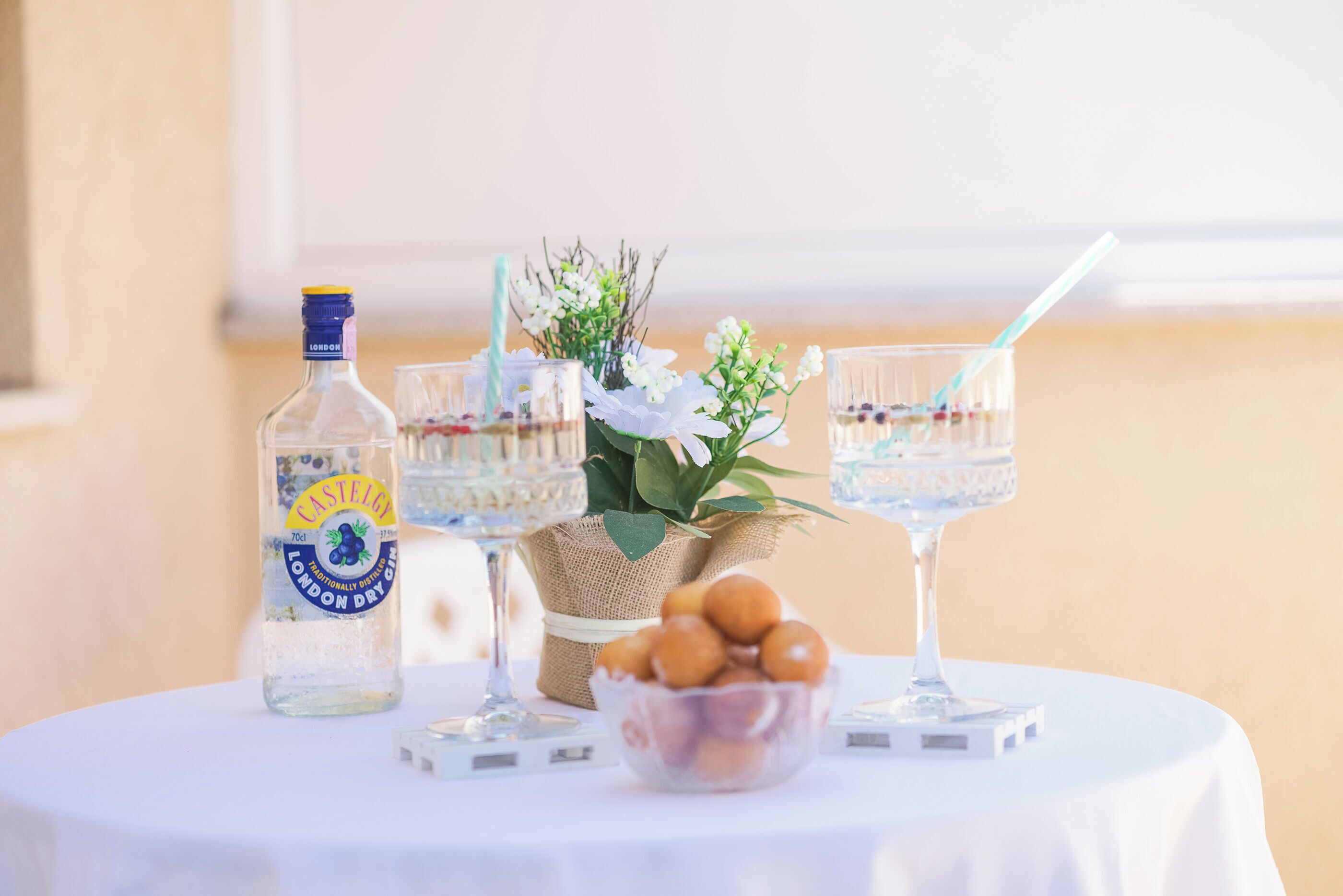 A table with a bottle of London Dry Gin, two glasses with straws, a bowl of round pastries, and a small plant.