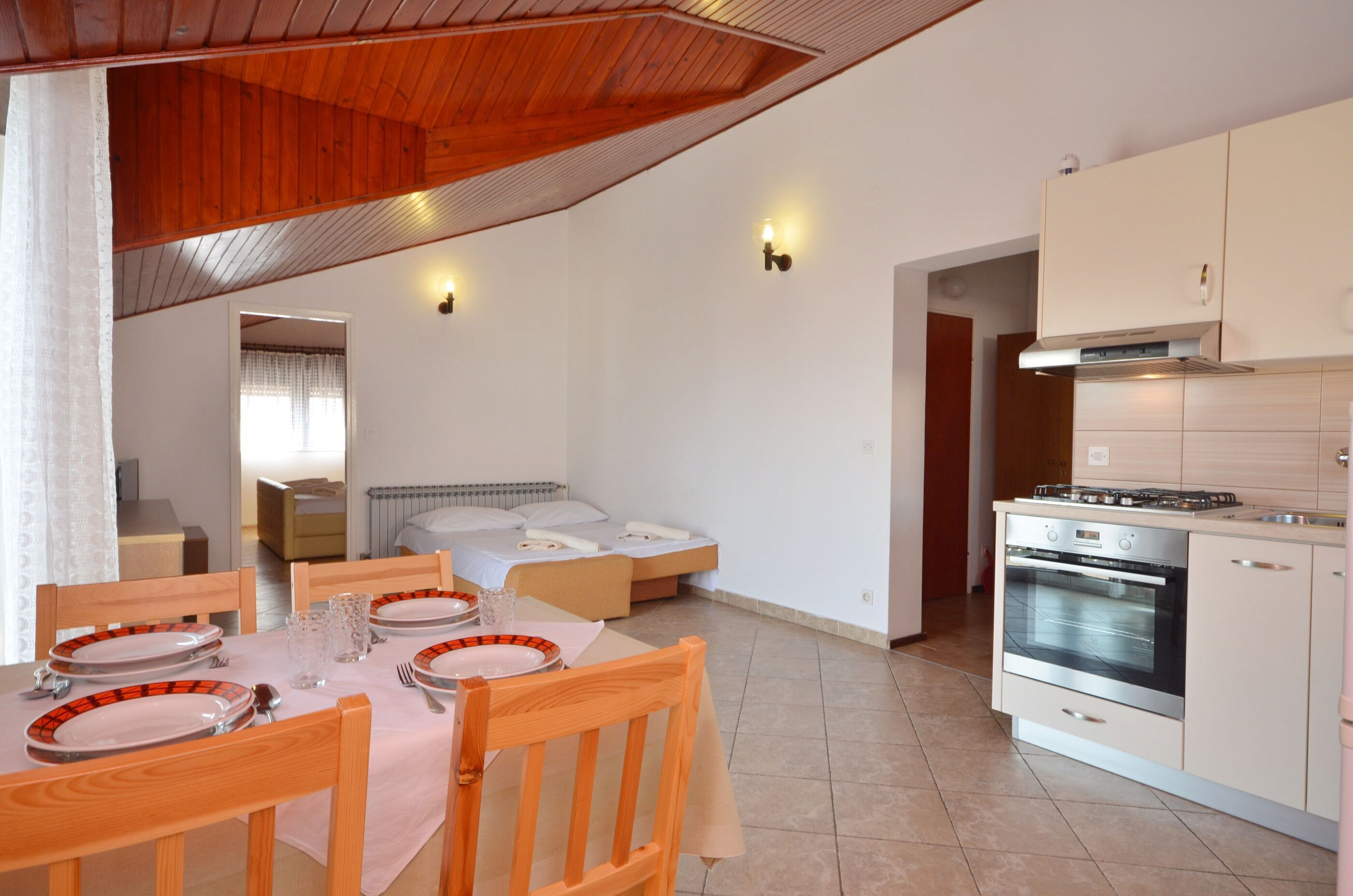 A compact kitchen with a stove, oven, and white cabinets. There is a dining table with chairs and a bed in the background.