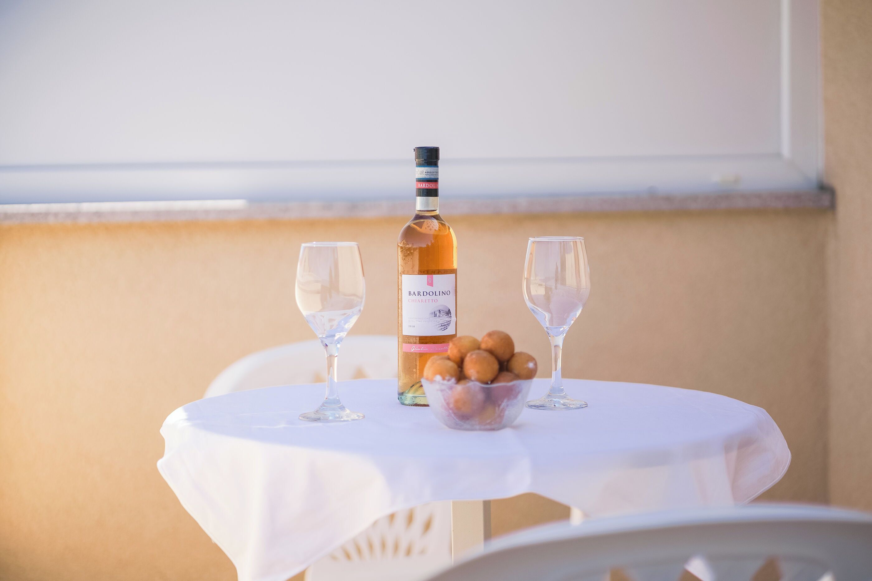 A table with a bottle of Bardolino Chianti, two empty wine glasses, and a bowl of snacks on a white tablecloth.