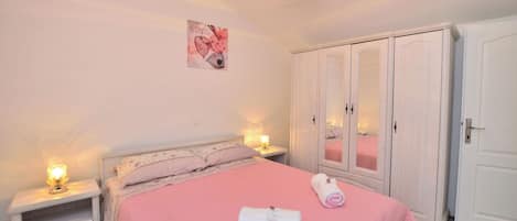 Quarto com colcha rosa, guarda-roupa branco e abajures nas laterais.
