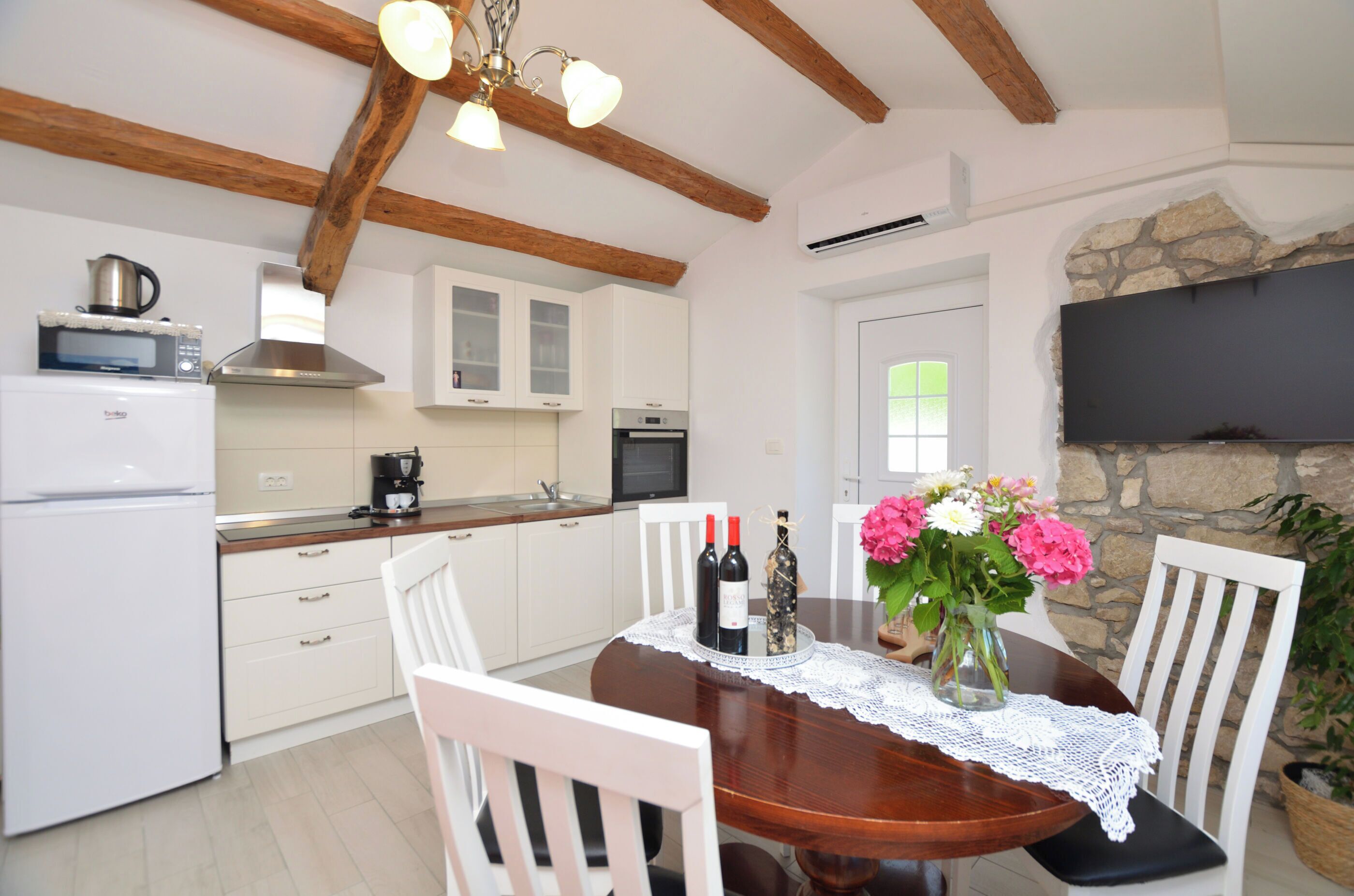 Een moderne keuken met witte kasten, een houten plafond en een eettafel met stoelen. Er hangt een televisie aan de muur en er staat een potplant aan de rechterkant.
