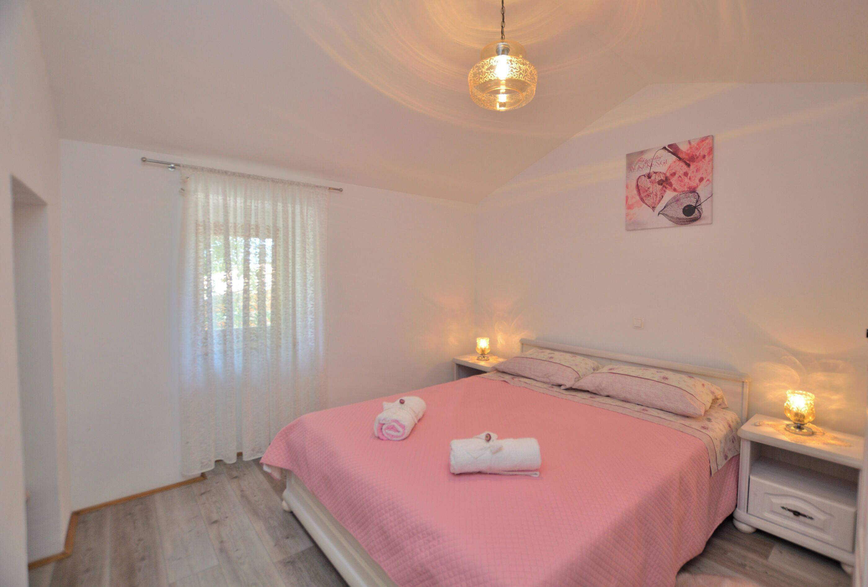 Ein Schlafzimmer mit rosa Bettdecke, zwei aufgerollten Handtüchern, einem Wandbild und einer Pendelleuchte.
